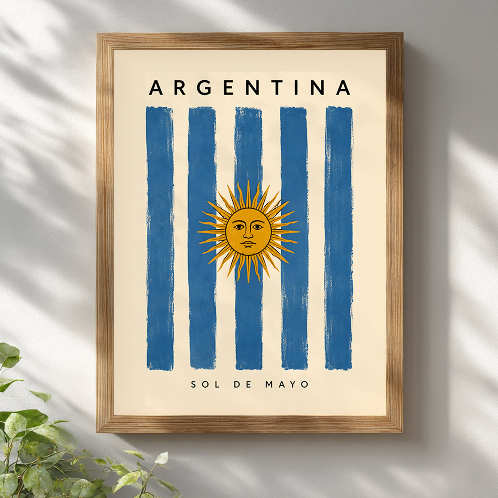 Argentina flag poster displayed in a warm wooden frame against a bright white wall with soft leafy shadows. The artwork features blue and cream stripes, the golden Sun of May, Argentina lettering and small Sol de Mayo text. The natural frame and light interior styling complement this vintage-inspired Argentine travel print.
