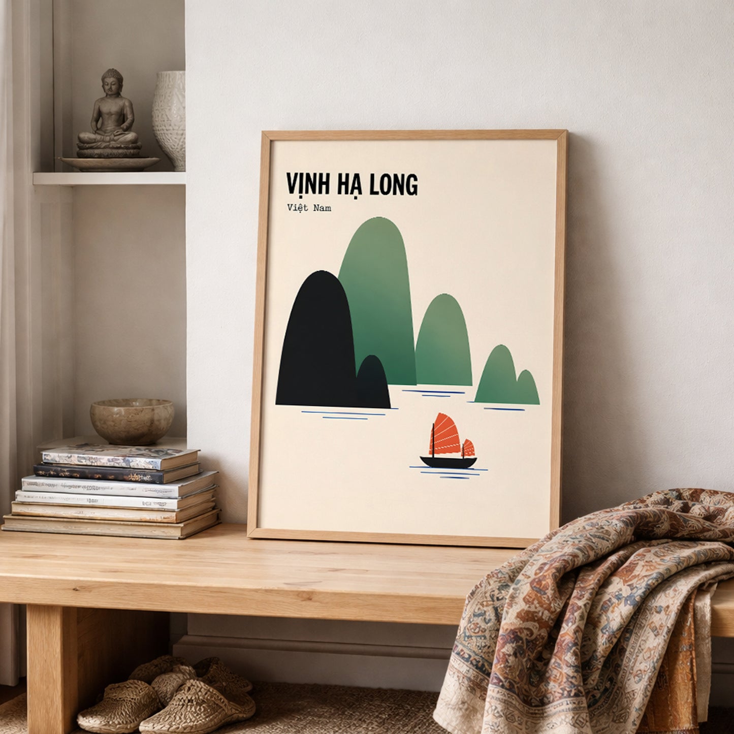 Vịnh Hạ Long travel poster in a light wooden frame, styled on a shelf in a soft neutral interior. The cream print features dark navy and green Ha Long Bay limestone islands with a small red sailboat, placed beside stacked books and natural décor with a textured patterned throw draped across the foreground.