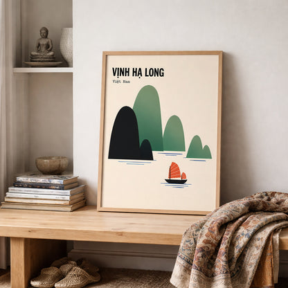 Vịnh Hạ Long travel poster in a light wooden frame, styled on a shelf in a soft neutral interior. The cream print features dark navy and green Ha Long Bay limestone islands with a small red sailboat, placed beside stacked books and natural décor with a textured patterned throw draped across the foreground.