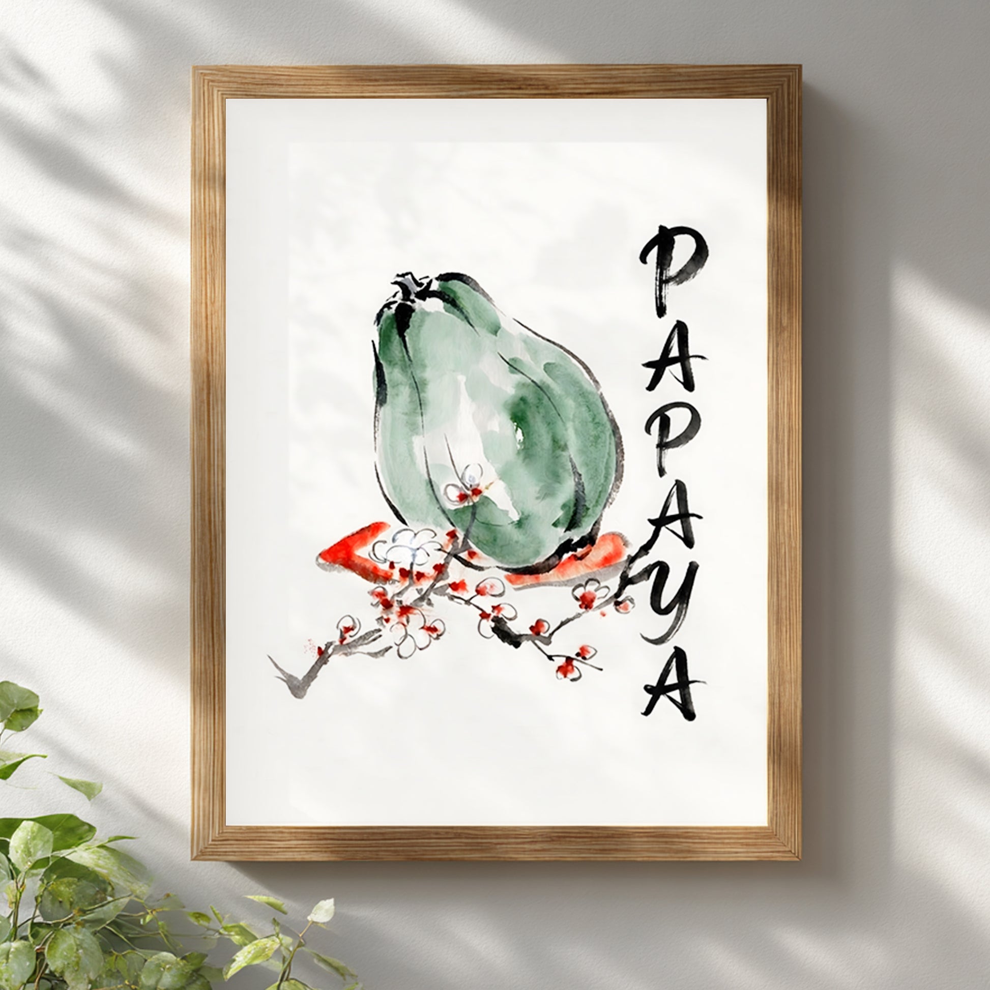 Framed papaya wall art in a light wood frame hanging on a white wall with soft natural shadows. The print features a green papaya painted in loose watercolour and black ink, a red and white blossom branch, and vertical PAPAYA text. Clean tropical fruit poster for kitchen wall art, dining room decor, botanical interiors, and modern minimalist home styling.
