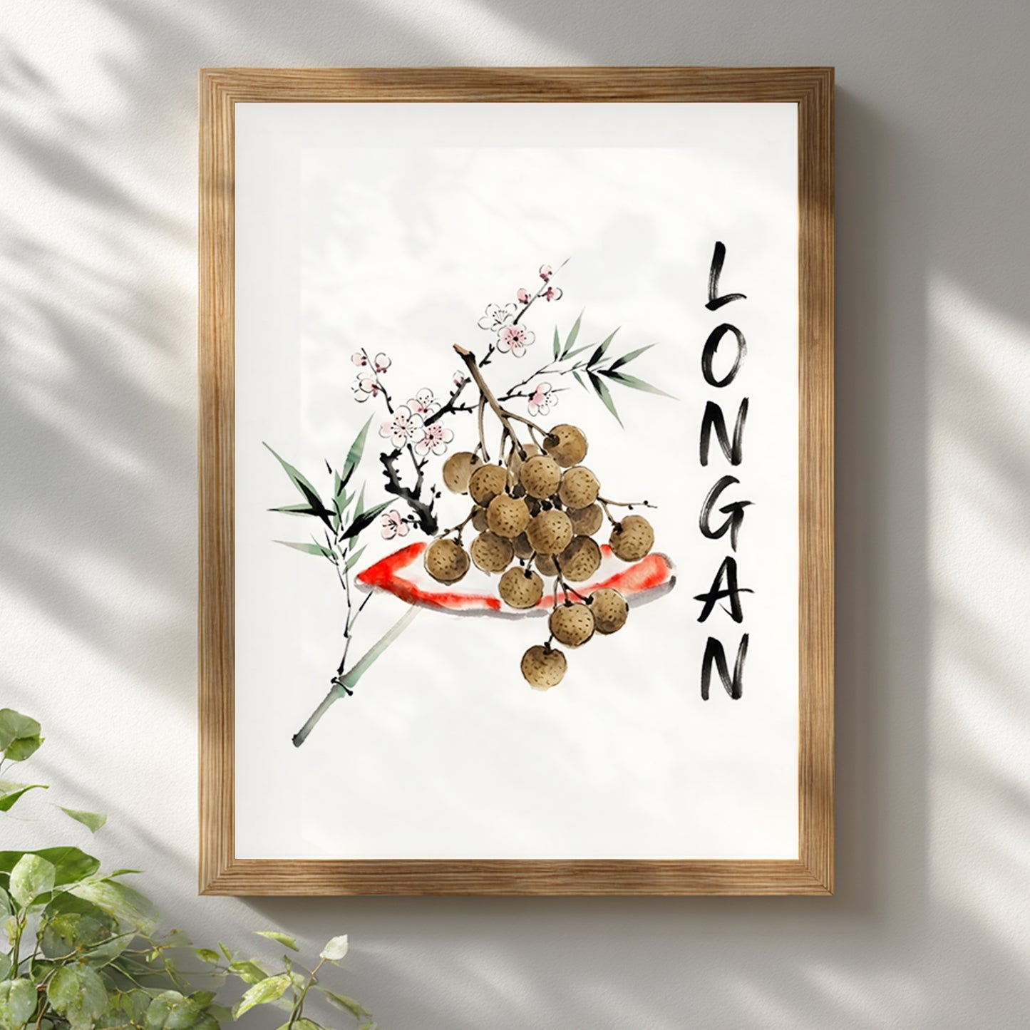 Wooden framed Thai longan watercolour print hanging on a white wall with soft natural shadows. The artwork shows brown longan fruit, green bamboo-style leaves, pale blossoms, red brushwork and vertical black lettering, styled as minimalist Southeast Asia inspired wall art for calm home decor.