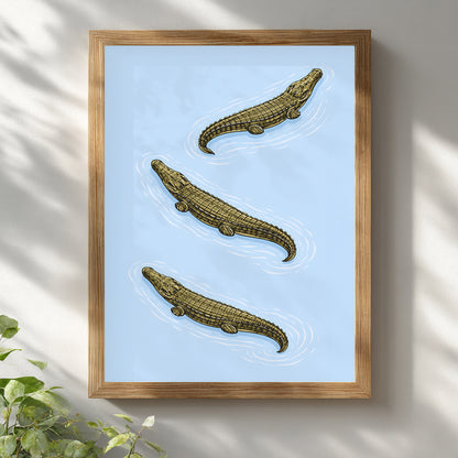 Triple Estuarine Crocodiles art print presented in a light wooden frame against a white wall. Three olive-toned saltwater crocodiles appear diagonally across a soft blue background, rendered in vintage zoological illustration style. A balanced reptile wall art piece suited to study rooms, home offices and modern natural history interiors.