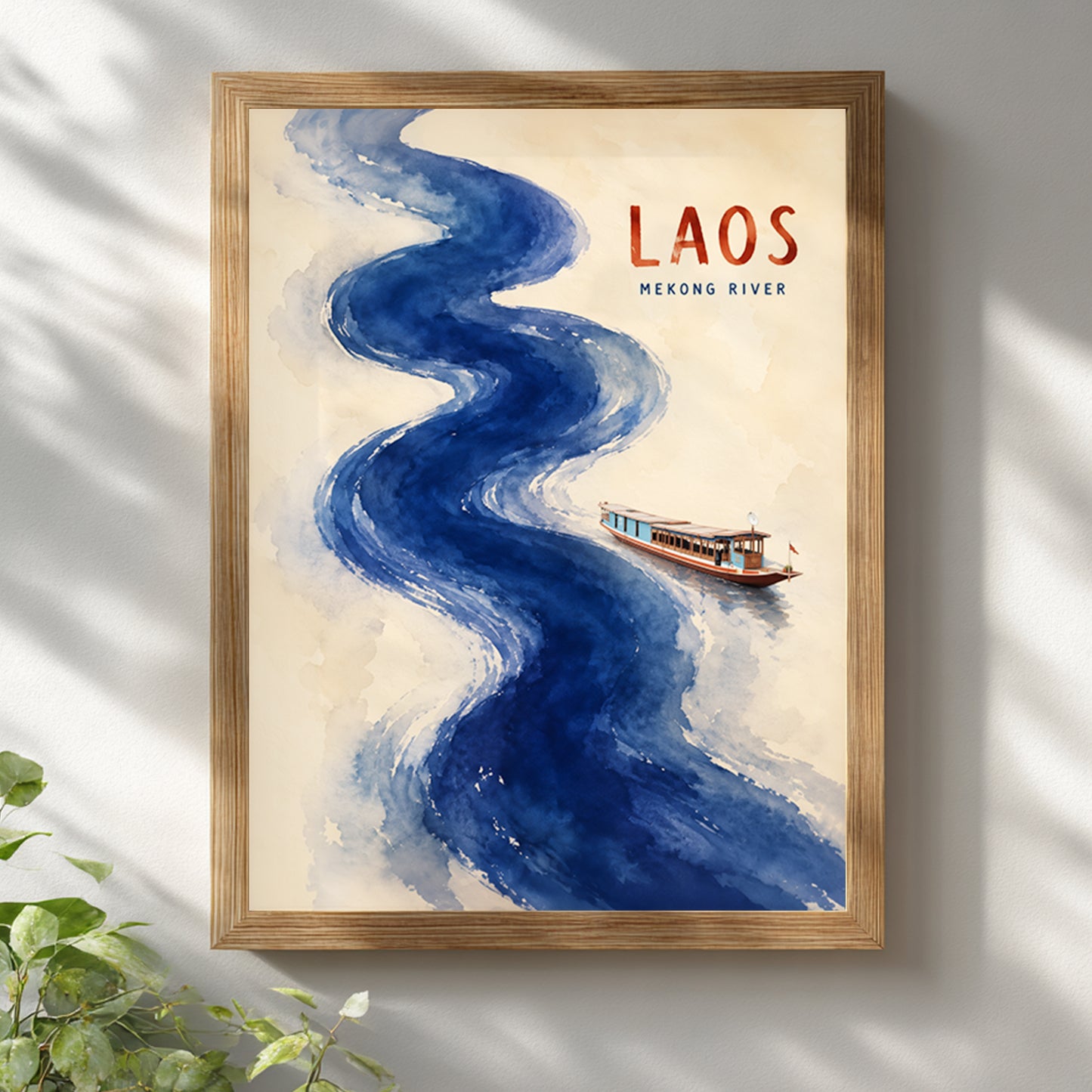 Laos Mekong River travel poster displayed in a light wooden frame on a bright white wall. The artwork shows a bold winding blue watercolour river, a small traditional boat and red LAOS lettering on a warm cream background. Soft daylight and green leaves create a fresh, natural setting for this Southeast Asian wall art.